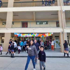 Fotografía de un estudiante junto a su apoderado en el patio del Colegio Integrado de Talca. En el fondo un grupo de estudiantes, docentes y funcionarios con globos de colores en el techo inflados con helio.